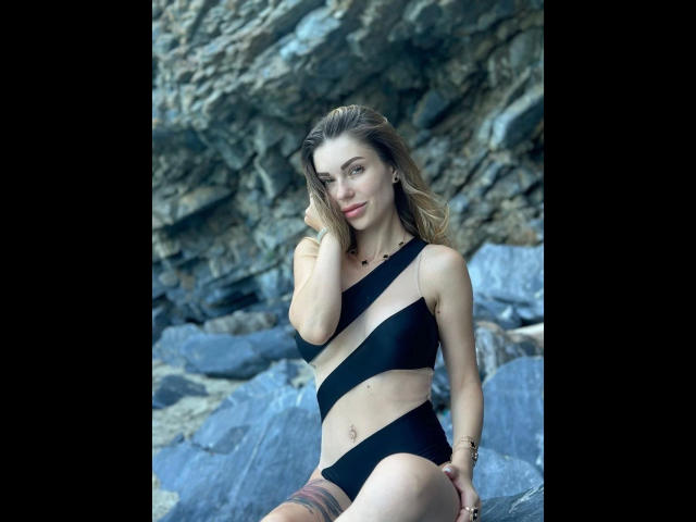 BerlinerGirl89-hot with blonde hair, black one-piece swimsuit, showing her tits and ass, sitting on rocks, hand in hair, smiling, tattoos on thigh, and gold bracelet.