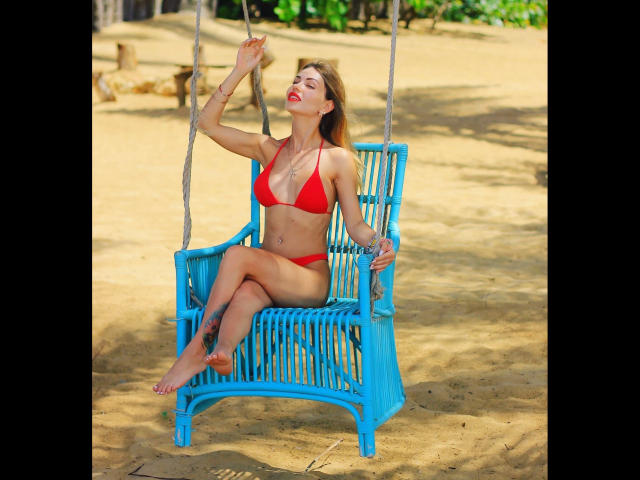 BerlinerGirl89-hot sits on a blue wooden swing, wearing a red bikini, with hands up, eyes closed, and red lipstick, showing her tits and pussy. She's on sandy ground.