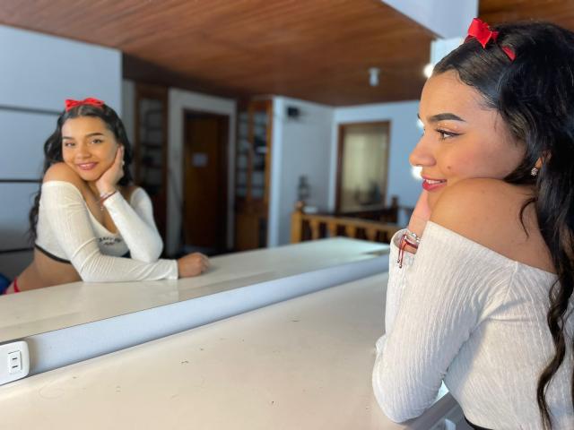 TaraConis, met donker haar, een rode strik, een witte top met open schouders, leunt tegen een toonbank, glimlacht naar haar spiegelbeeld, waarbij haar blote schouders en rode oorbellen zichtbaar zijn, in een kamer met houten lambrisering..