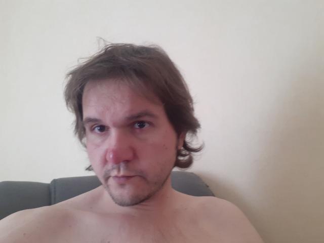 NectarineFad has a slightly red nose, brown wavy hair, light skin, and a small goatee, with a neutral expression, shirtless, and sitting against a plain wall.