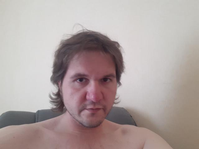 NectarineFad has a chubby face, messy brown hair, and a slight beard, with a serious expression, shirtless, and a pink nose, sitting against a plain wall.