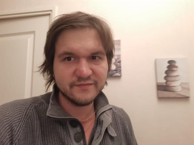 NectarineFad has light brown, shaggy hair, a light beard, and is wearing a gray, textured, button-up shirt. NectarineFad's face shows a slight, confident smile.