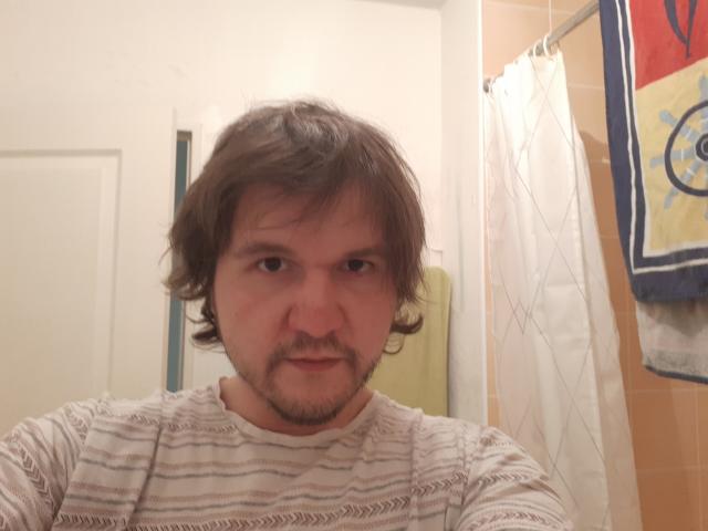 NectarineFad has medium-length brown hair, a light beard, and is wearing a striped white shirt. He looks serious with a slight smirk. Bathroom background.