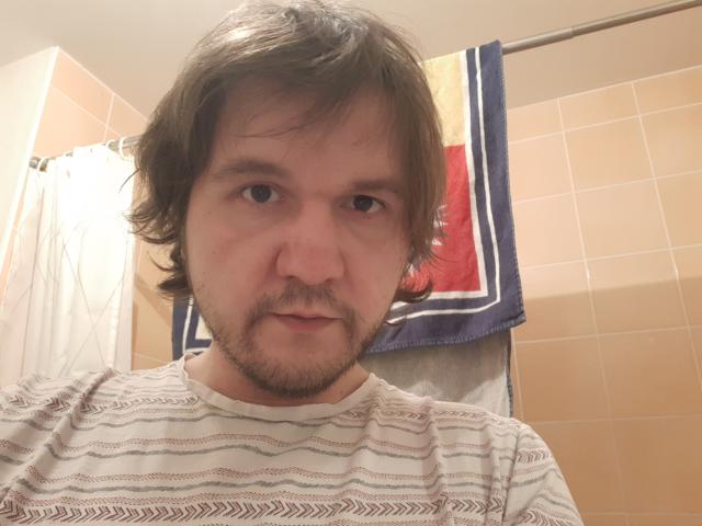 NectarineFad has brown, wavy hair, a beard, and a serious look. He's wearing a striped shirt, and there's a blue towel with a red and yellow pattern behind him.