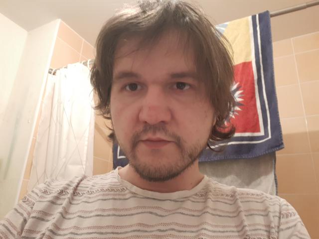 NectarineFad has a serious face, brown shaggy hair, a small beard, and is wearing a striped shirt. His nose is slightly red and he looks tired.