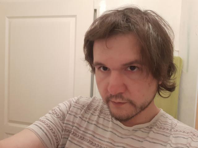 NectarineFad with messy brown hair, small beard, and striped shirt, giving a serious look, showing slightly red eyes, wearing a white shirt with brown patterns.