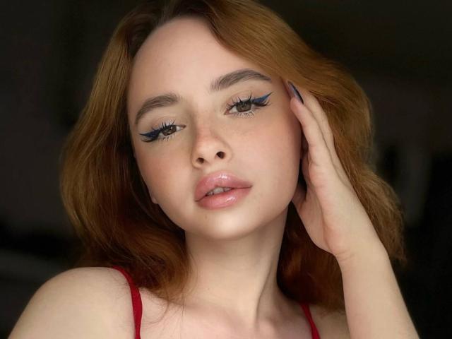 EllaAndDani has red hair, blue eyeliner, and glossy pink lips, with a hand touching her face, wearing a red top, in a soft-lit, slightly blurred photo.
