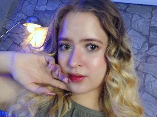 Emily Kane tiene el cabello rubio y rizado, ojos verdes y lápiz labial rojo. Está tocándose la mejilla con la mano derecha y lleva una blusa verde. Sus pezones no son visibles..