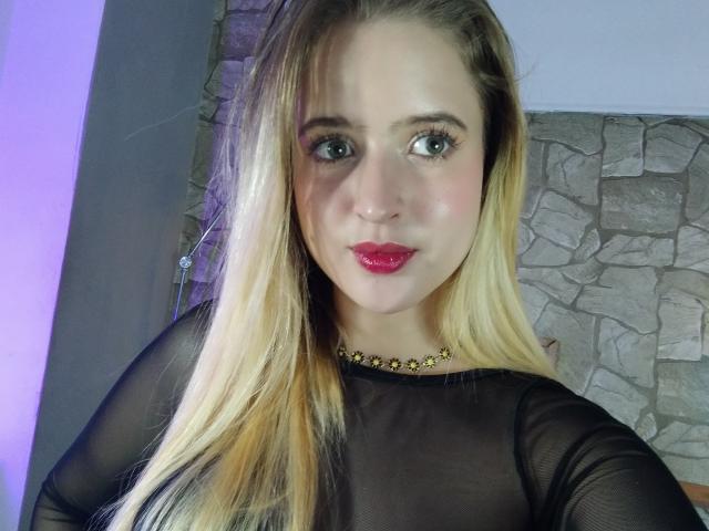 EmilyKane with blonde hair, blue eyes, and red lipstick, wearing a black sheer top and a gold star necklace, has a cute face and is looking at the camera.