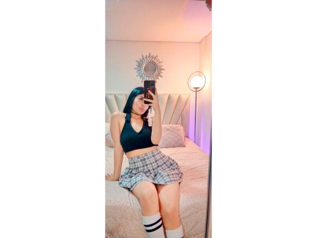 EmilyJensenn with blue hair, black sports bra, plaid skirt, white knee socks, taking a mirror selfie, showing her ass, sitting on a bed, holding a phone.