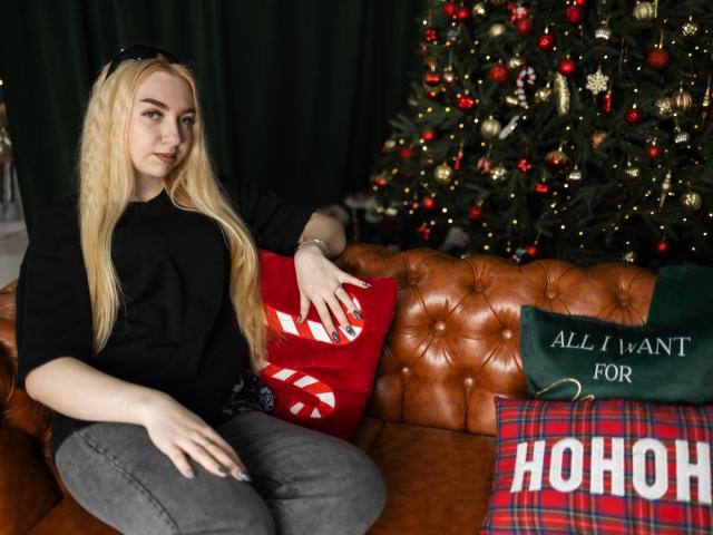 BeatriceFly, une blonde aux longs cheveux, vêtue d'une chemise noire et d'un pantalon gris, est assise sur un canapé en cuir marron, les bras reposant sur des coussins de Noël rouges et verts, à côté d'un sapin décoré..