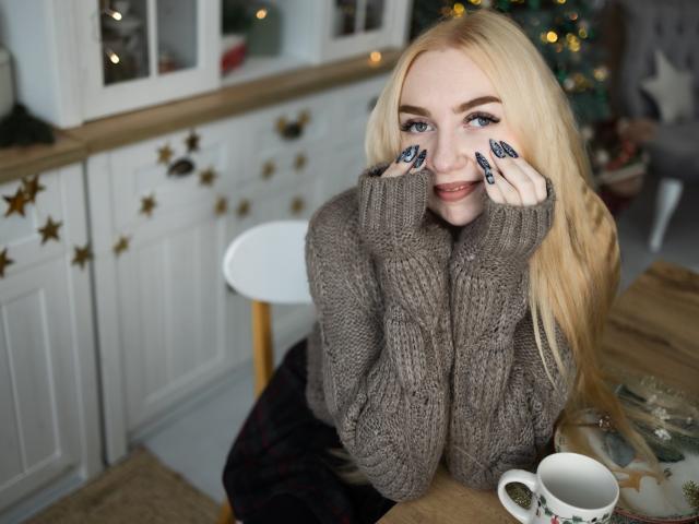 Blonde BeatriceFly with long hair, blue eyes, and dark nail polish, wearing a grey knit sweater, resting hands on cheeks, smiling, sitting at a wooden table with a white cup.