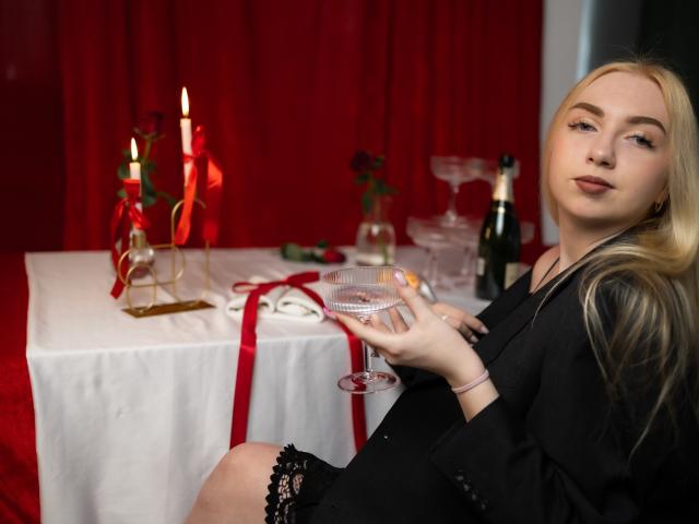 Blonde BeatriceFly with tits in black lingerie, holding a glass, sitting by a red-clothed table with candles and a bottle, looking at the camera with a slight smile.