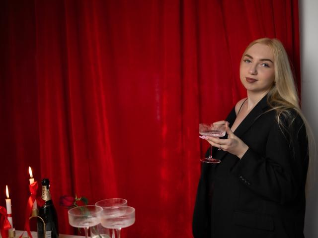 Beatrice, una mujer con cabello rubio largo, vestida con un blazer negro, sosteniendo una copa de vino rosado, de pie junto a una mesa con velas y vasos de agua..