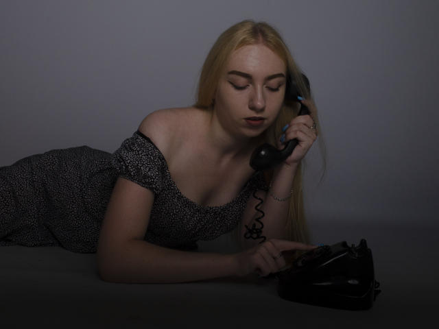 BeatriceFly with blonde hair, off-shoulder black and white dress, lying on floor, holding old black phone to ear, looking down, fingers on receiver, gray background, subtle lighting.