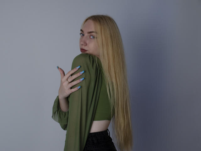 Beatrice, une femme aux longs cheveux blonds, portant un haut vert, un pantalon noir, avec des ongles bleus, regardant par-dessus son épaule, tenant le haut vert, montrant son dos et une partie de ses fesses..