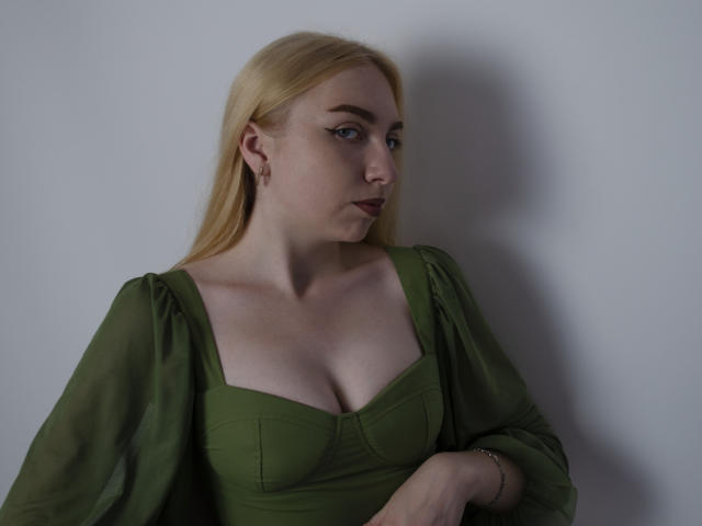 BeatriceFly with blonde hair, green dress, and puffy sleeves, showing her tits in a photo, looking to the side with a serious expression, white background.