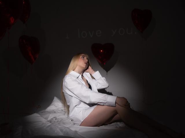 BeatriceFly with long blonde hair, sitting on a bed, wearing an oversized white shirt, exposing her bare ass, surrounded by red heart balloons, in dim lighting.