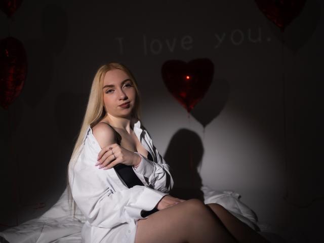 Blonde BeatriceFly mit langen Haaren, die ein offenes weißes Hemd trägt und einen schwarzen BH zeigt, sitzt auf einem Bett. Im dunklen Raum ist der Schatten eines roten Herzens und die Worte "Ich liebe dich" zu sehen..