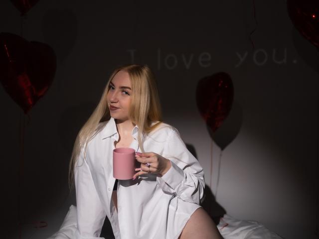 Blonde BeatriceFly mit langen Haaren, die ein offenes weißes Hemd trägt, eine rosa Tasse hält und auf einem Bett sitzt, wobei ihr nackter Hintern zu sehen ist, in einem schwach beleuchteten Raum..