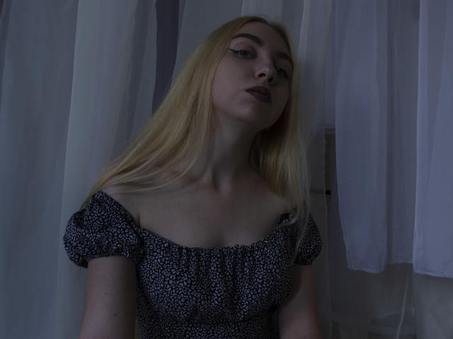 Beatrice, mit blonden Haaren, trägt ein schwarzes und weißes Oberteil mit Blumenmuster und einem Schulterteil, hat ein leicht schmollendes Gesicht und dunklen Lippenstift, bei gedämpftem Licht. Ihre Brüste sind klein..