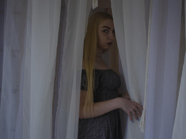 Beatrice, con el pelo rubio largo, lápiz labial oscuro y uñas azules, está de pie frente a cortinas blancas, vestida con un vestido negro con lunares blancos, mirando hacia un lado..
