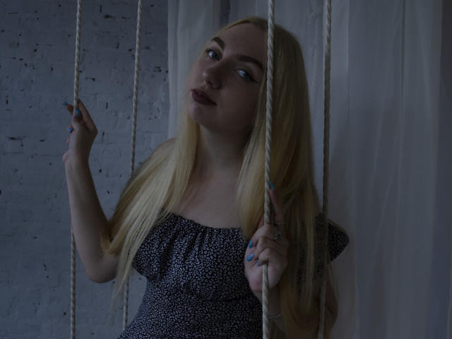 BeatriceFly with long blonde hair, wearing a black speckled dress, holding two ropes on a swing, looking slightly up, with dark lipstick and blue nail polish.