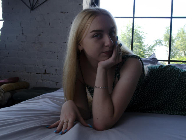 Blond BeatriceFly with blue nails, wearing a green polka-dot dress, leans on a bed, hand under chin, looking thoughtful, sunlight through window, white brick wall background.
