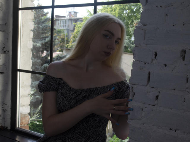 BeatriceFly with long blonde hair, off-shoulder black dress with white dots, blue nails, holding her tits, standing by a white brick wall, looking outside a window.