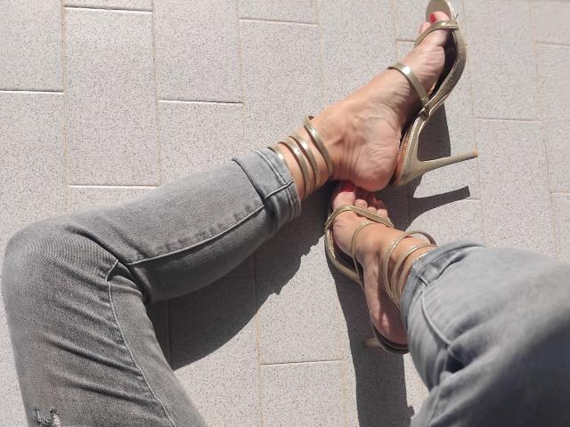 Rosyta's feet in gold high heels, with her grey jeans pulled down to expose her toes and ankles. Her skin is pale, and she's standing on a tiled floor.