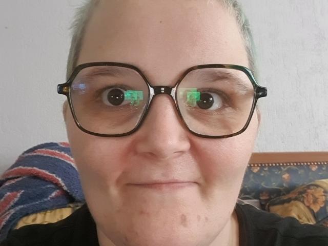 Lya69x hat ein rundes Gesicht mit Brille, grüne Reflexionen in den Augen, ein leichtes Lächeln, helle Haut, kleine Pickel am Kinn und kurze, graue Haare. Im Hintergrund sind gestreifte und gemusterte Kleidungsstücke zu sehen..