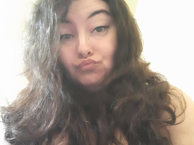 BebeMilfForU with pouty lips, dark wavy hair, and black eyeliner, giving a playful kiss to the camera, her cute face centered, soft light on her fair skin.