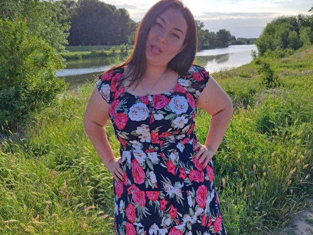 BebeMilfForU with hands on hips, wearing a floral dress, sticking out tongue, has pink flowers on black dress, medium tits, brown hair, standing in green grassy area.