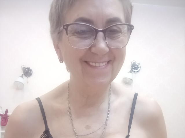 MadamMargo with short brown hair, glasses, smiling, wearing a black lace top, necklace, and earrings, showing small tits and a cute face.
