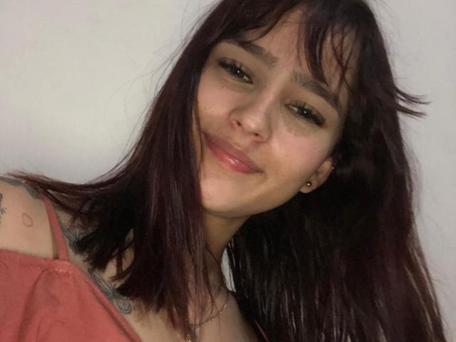 MelodyMansson with dark red hair, small nose ring, smiling, wearing pink top, tattoo on shoulder, brown eyes, slight blush, ear stud, relaxed, upper body shot.
