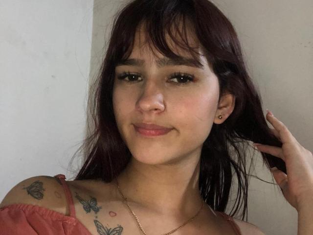 MelodyMansson has dark red hair, small nose, and light skin. She's smiling with a hand in her hair, showing butterfly tattoos on her shoulder and chest.