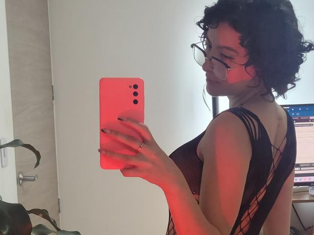 GabrielaMoore with curly black hair, glasses, and red lighting, wearing a black mesh top, taking a mirror selfie with a red phone, showing her back and ass.