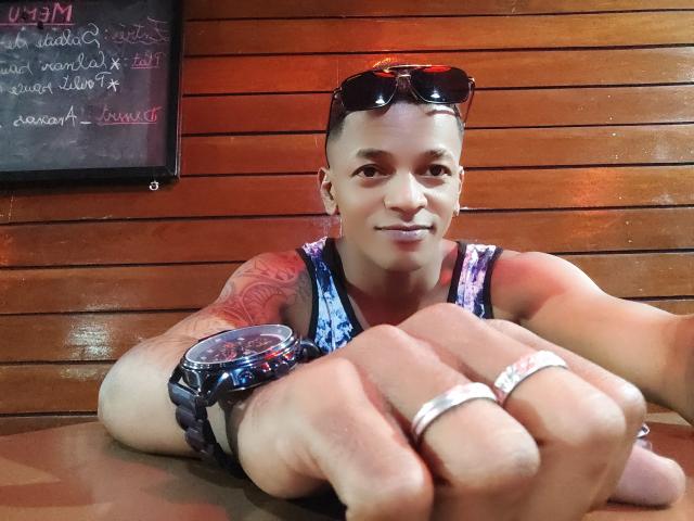 SexyCraigX, muscular, tan-skinned, with short hair and tattoos, wears a blue patterned tank top, black sunglasses on head, rings on fingers, and a watch, smiling at camera.