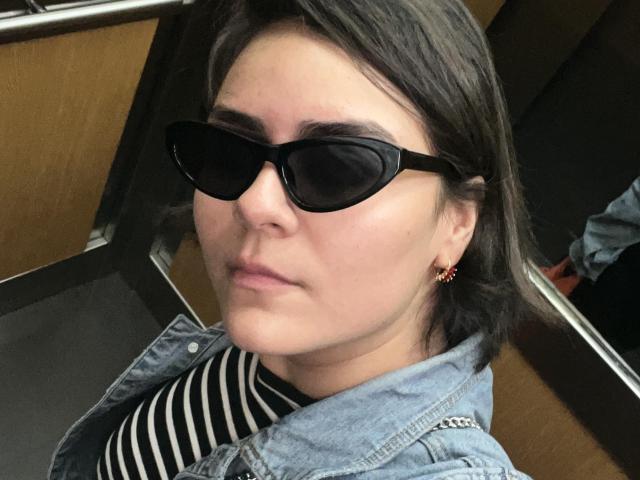 Isabela Cooper, con gafas de sol negras, cabello corto y oscuro, vistiendo una chaqueta vaquera sobre una camisa a rayas, un pequeño pendiente de flor roja, con una expresión ligeramente fruncida, en un ambiente interior con poca luz..