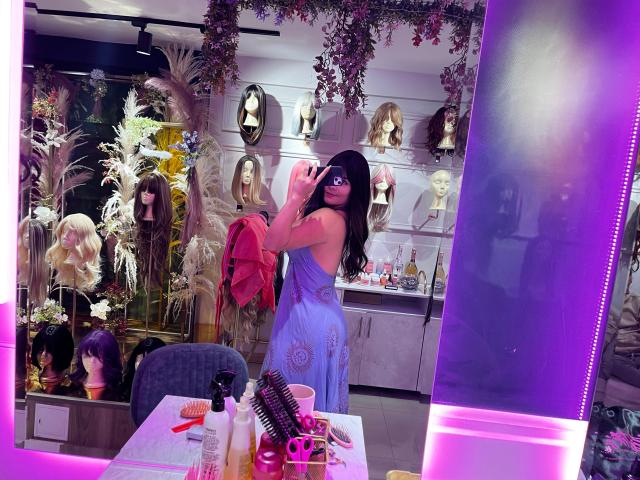 Isabela Cooper mit langem, schwarzem Haar, die ein violettes Kleid trägt und in einem Friseursalon ein Selfie vor einem Spiegel macht. Sie hält ein rosa Handy und ist von Dummys und Haarpflegeprodukten umgeben..
