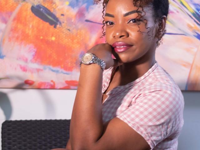 DannaBanks, avec ses cheveux bouclés foncés, portant une chemise rose à carreaux, a une montre argentée à son poignet, et repose son menton sur sa main, souriant avec du rouge à lèvres rose..
