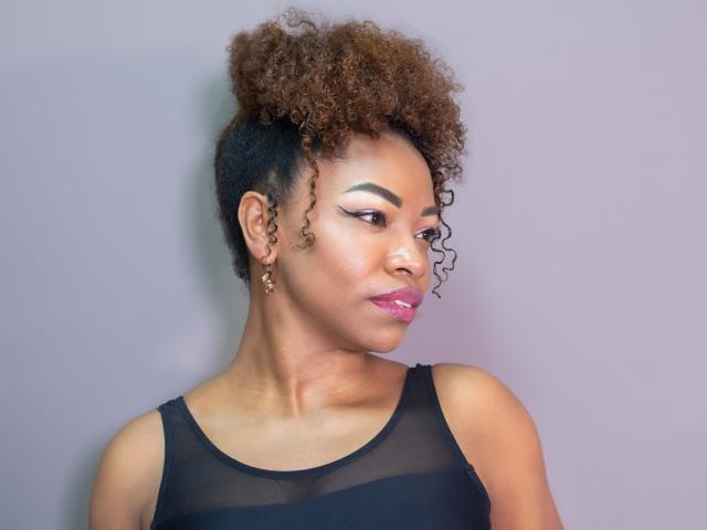 DannaBanks heeft krullend bruin haar, donkere huid en roze lippenstift. Ze draagt een zwarte, doorschijnende top, kijkt naar de zijkant met een lichte glimlach en heeft gouden oorbellen..