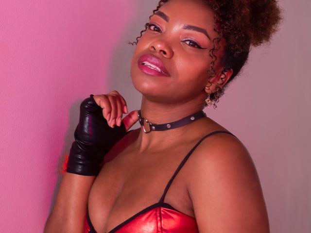 DannaBanks met krullend bruin haar, roze lippenstift, een zwarte choker, een rode leren top en een zwarte handschoen zonder vingers, kijkt omhoog met een lichte glimlach en houdt haar sleutelbeen vast..
