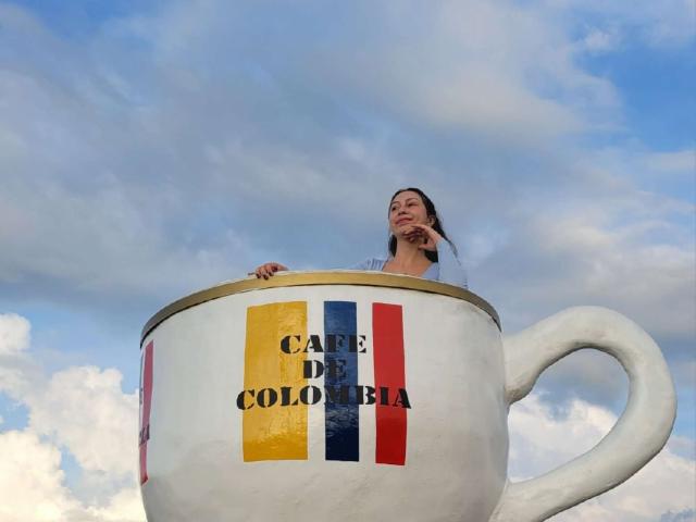 Emma Wells dentro de una taza blanca gigante con rayas rojas, amarillas y azules, mirando hacia abajo con una ligera sonrisa, cielo nublado arriba, el asa de la taza está a la derecha..
