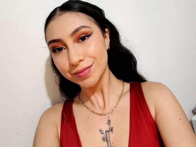 Emma Wells, com cabelos longos e pretos, batom rosa e vestido vermelho, exibindo seios pequenos e uma tatuagem de cruz acima do colo, usando uma corrente de ouro, sorrindo para a câmera..