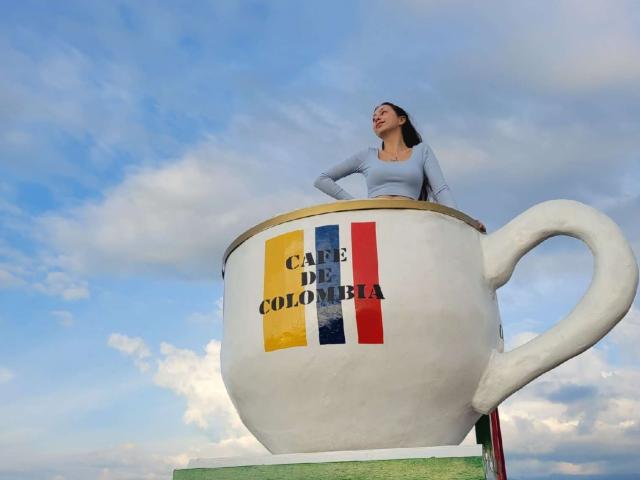 Emma Wells, con sus glúteos apoyados en una taza de té blanca gigante, vistiendo una blusa azul claro, de pie frente a un cielo azul nublado. La taza de té tiene un patrón de rayas verticales de colores..