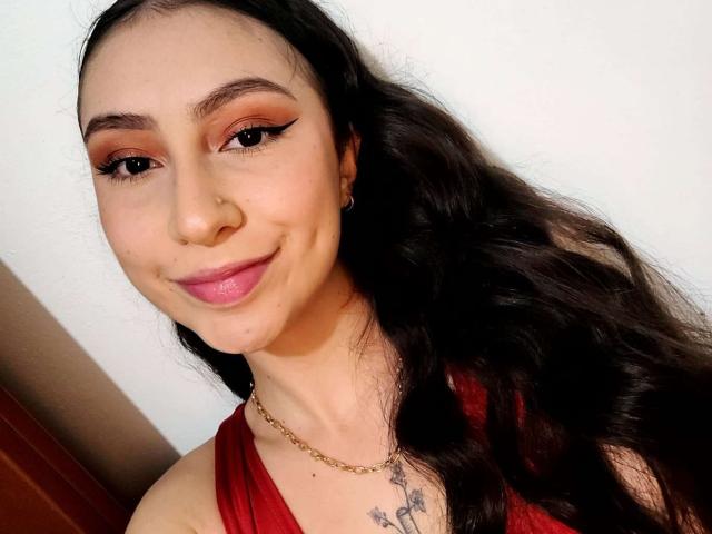 Emma Wells tem cabelos longos e pretos, batom rosa e sombra laranja. Ela está vestindo uma blusa vermelha, um colar dourado e tem um pequeno piercing no nariz, com um sorriso adorável..