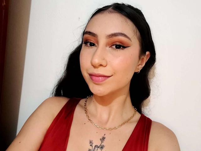 EmmaWells with long black hair, red dress, gold chain, and winged eyeliner, smiling at the camera, showing a small tattoo on her chest with two stars.
