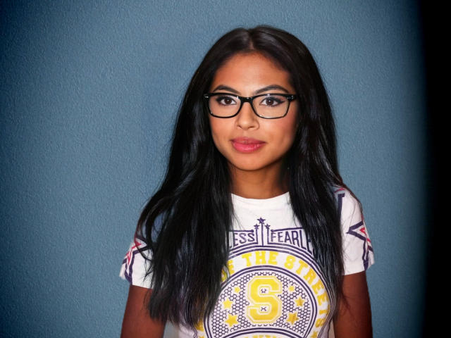 SoniaLegacy has long black hair, glasses, and a cute face with a slight smile. She's wearing a white shirt with a yellow and purple design. Boobs are not visible.