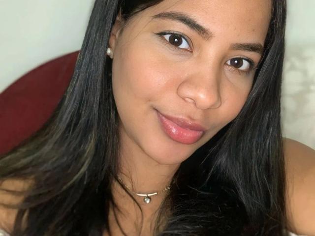 ThaisNunes-hot tem pele marrom lisa, cabelo preto longo, olhos castanhos, lábios rosados, e está usando um colar com um pequeno pingente de pérola. Seu rosto adorável está ligeiramente inclinado..